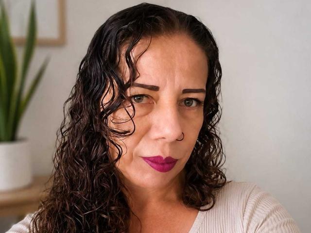 LaylaMilf has curly brown hair, a nose ring, and purple lipstick. She's wearing a beige top with her eyebrows slightly raised, looking directly at the camera.