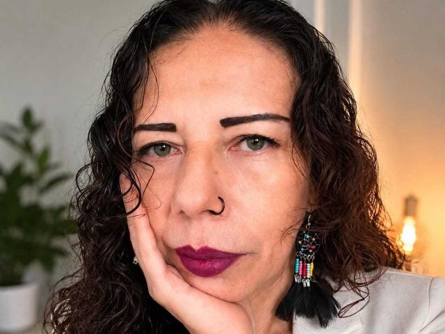 LaylaMilf has curly brown hair, green eyes, thick eyebrows, a nose ring, purple lipstick, and black tassel earrings. She's resting her face on her hand, looking serious.