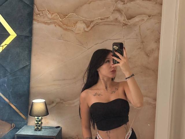 NikkiGoldX with long black hair, black strapless top, tattoos on chest, holding phone, mirror selfie, lamp on left, marble wall, NikkiGoldX's tits visible, NikkiGoldX's ass not visible.