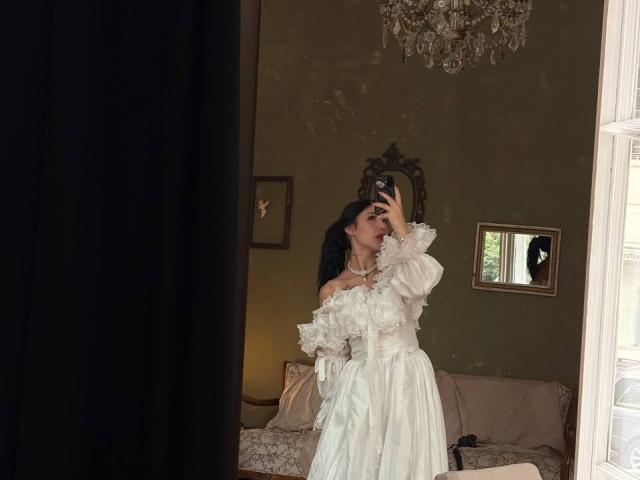 NikkiGoldX in a white off-shoulder dress with ruffles, holding a phone, taking a mirror selfie, showing her tits, with long black hair, in a dimly lit room.