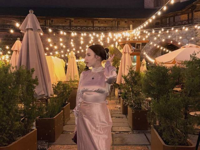 NikkiGoldX with pale pink satin dress, long sleeves, standing in outdoor area, dark hair in bun, right hand raised, surrounded by string lights, green plants, and beige umbrellas.