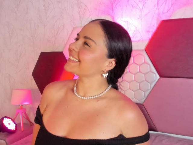 ArielleWilde with dark hair, off-shoulder black top, pearl necklace, and earrings, smiling, showing her tits, pink hexagon-patterned headboard, and pink lamp in background.