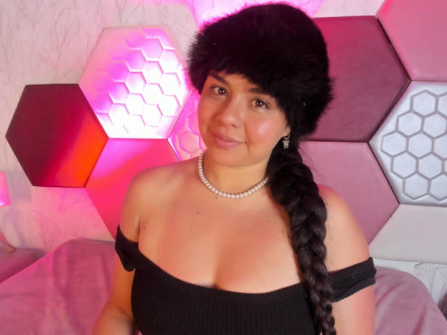 ArielleWilde with black off-shoulder top, pearl necklace, and fur hat, braided black hair, pink and white hexagonal wall, slight smile, medium tits, cute, bedroom.