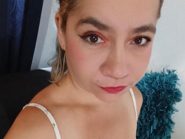 MsJustineBeaux has light skin, blonde hair in a ponytail, red lipstick, and subtle eyeliner. She's wearing a white spaghetti strap top, showing her small tits.