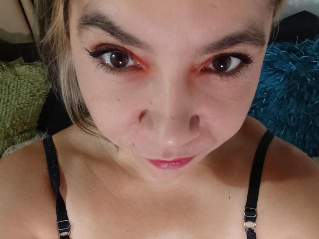 MsJustineBeaux's face with brown eyes, red eyeliner, and pink lipstick, wearing a black strap bra, looking up with a slight pout, showing her nose and subtle freckles.