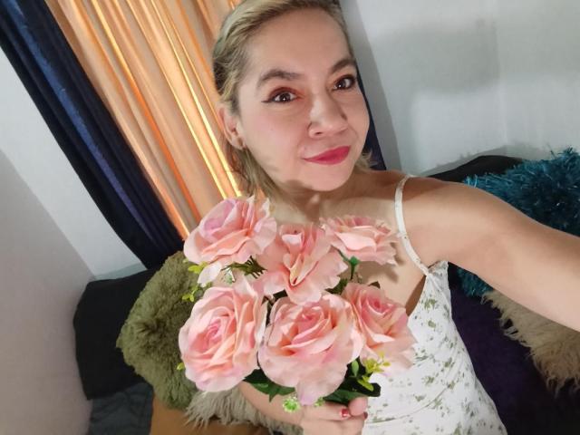 MsJustineBeaux with blonde hair, pink roses in hand, white floral dress, smiling, holding flowers to camera, sitting on couch with blue and green pillows.