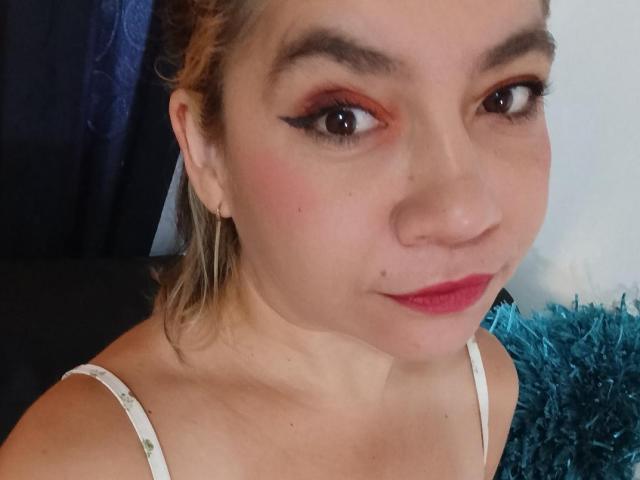 MsJustineBeaux's face, showing light skin, red eyeshadow, black eyeliner, pink lipstick, small nose, blonde hair in a ponytail, white spaghetti strap top, and a slight smile.