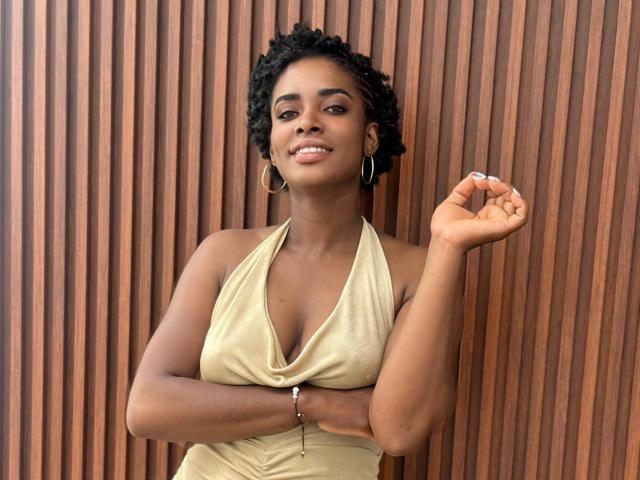 TiaraAlamo has dark skin, curly black hair, and is wearing a gold halter dress, showing her small tits and smiling with hoop earrings, holding a finger up.
