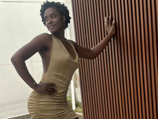 TiaraAlamo with dark skin, curly black hair, wearing a gold, ruched, side-cutout dress, leaning against a wooden wall, smiling, hand on hip, other hand on wall.