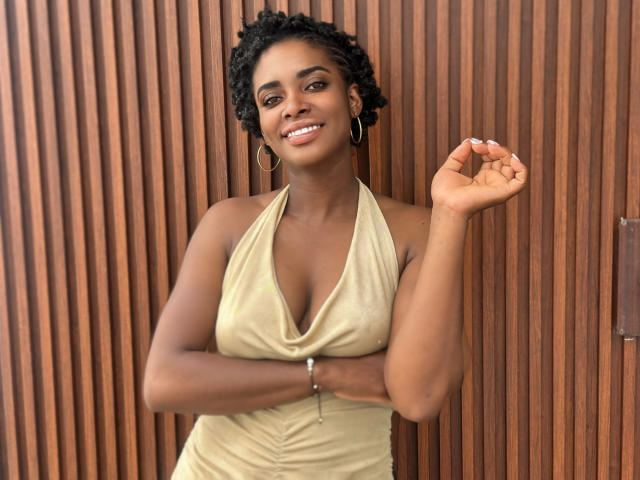 TiaraAlamo, dark-skinned with curly black hair, wearing a beige halter dress, smiling with her hand up, showing her white nails, and wearing gold hoop earrings.
