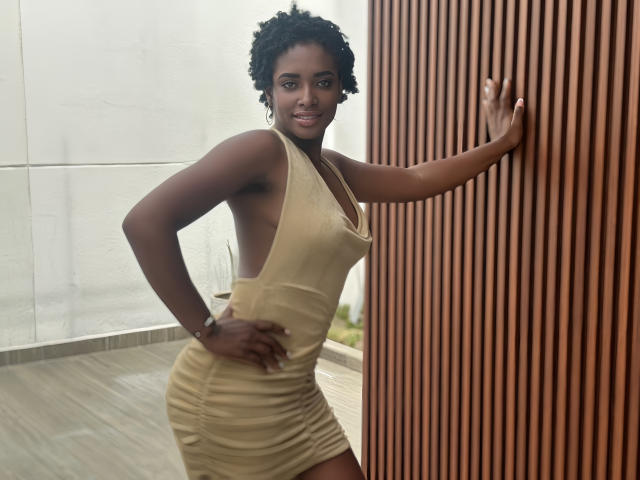 TiaraAlamo with dark skin, curly hair, and a smile, wearing a tight, side-cut, yellow dress, standing against a wooden wall, hand on it, other hand on hip.