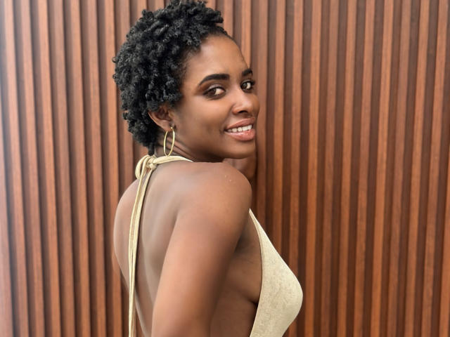 TiaraAlamo with dark brown skin, curly black hair, gold hoop earrings, and a backless, tan dress, smiling over her shoulder, showing her side boob.