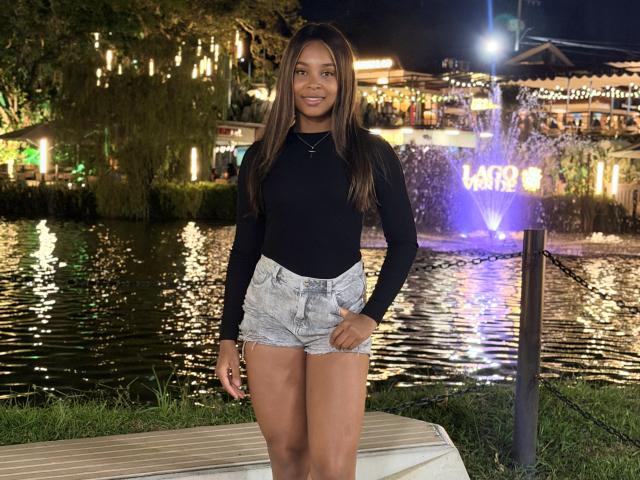 Emma, com cabelos castanhos longos, blusa de manga comprida preta e shorts jeans de cintura alta, está parada perto de uma fonte de água, sorrindo, com a mão na coxa..