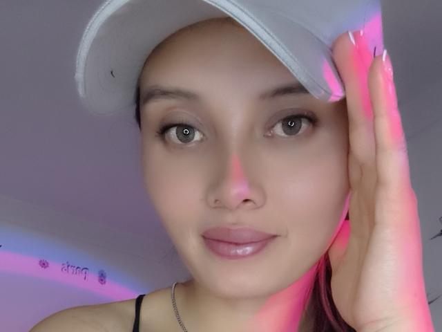 Ahllana with fair skin, brown eyes, and pink lips, wearing a gray cap, touching her forehead with pink-named nails, showing a subtle smile. She has small tits.