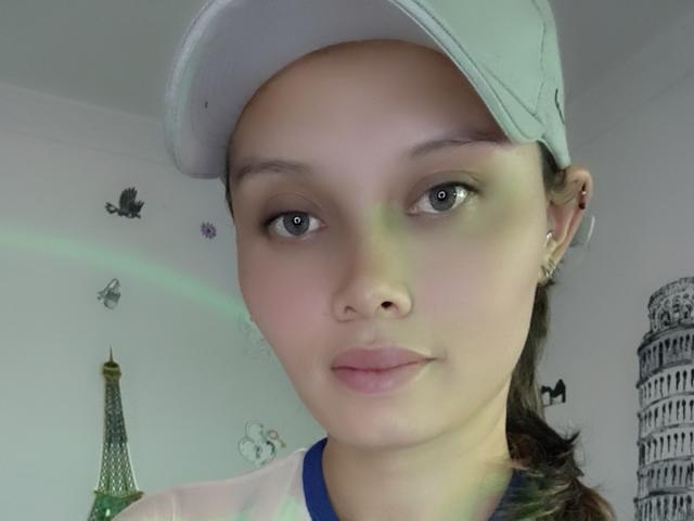 Ahllana with a neutral expression, wearing a grey cap, has light skin, green eyes, and pink lips, with small earrings, and brown hair in a ponytail. Her tits are not visible.