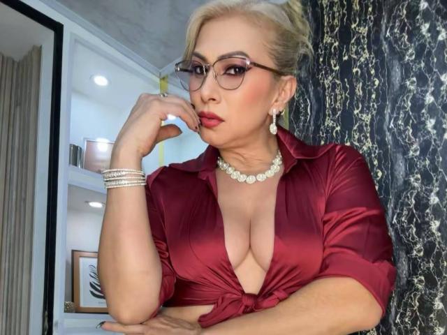 MisseScarleth with blonde hair, glasses, red shirt, deep cleavage, pearl necklace, pearl earrings, and pearl bracelet, resting chin on hand, looking serious.