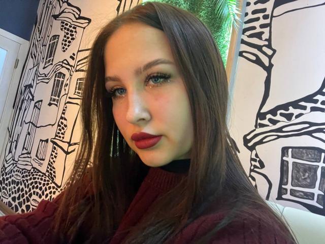 VilmaSwamy with long brown hair, red lipstick, and a maroon sweater, wearing green earrings, looking at the camera with a slight smile, blue eyes, and a cute face.