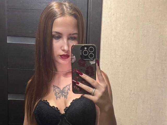 VilmaSwamy with long brown hair, red lipstick, black lace bra, butterfly tattoo, holding phone, showing off her tits, and red nails, wearing a silver necklace.