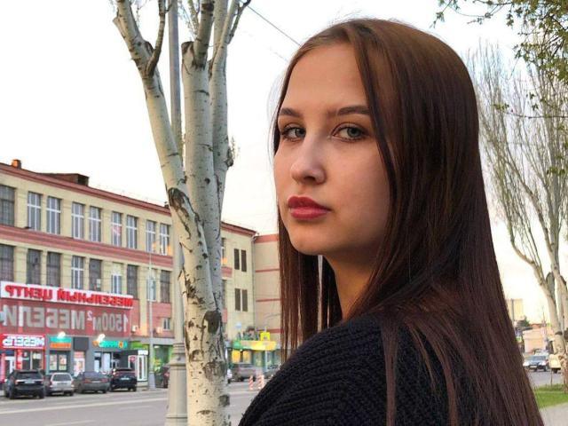 VilmaSwamy has long brown hair, green eyes, and red lipstick, wearing a black knit sweater, standing by a birch tree with a serious expression.