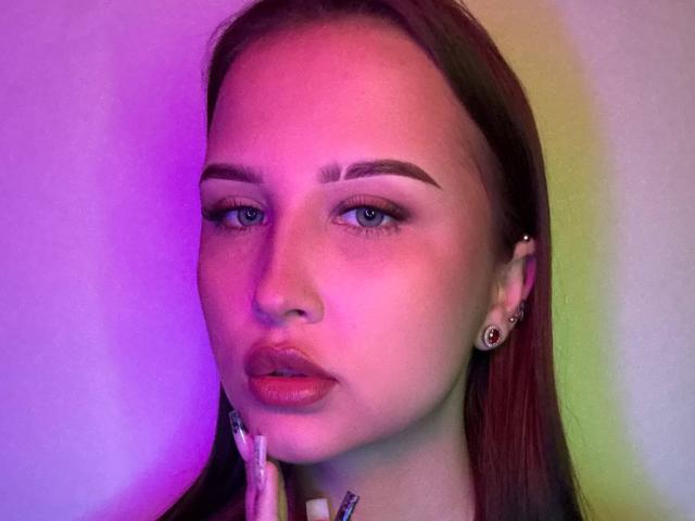 VilmaSwamy's face with pink and purple light, holding a makeup brush near her mouth, has brown hair, dark eyebrows, and pinkish lips, wearing small earrings.