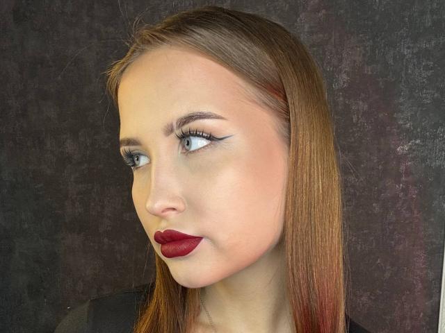 VilmaSwamy has pale skin, blue eyes, dark red lipstick, and long brown hair with red tips. She's wearing black eyeliner and looking to the side.