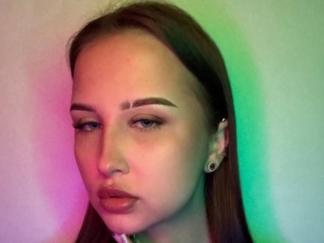VilmaSwamy has a serious face with pouty lips, brown hair, and green-blue eye makeup. She has ear piercings and a slightly asymmetrical face with pink-green lighting.