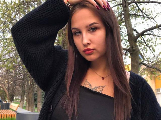 VilmaSwamy with long brown hair, black cardigan, red lipstick, and tattoo on chest, hand in hair, black top, red nails, necklace, and wristwatch, standing outdoors.
