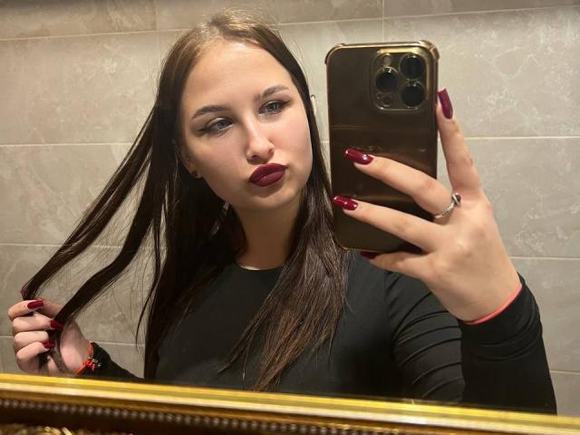 VilmaSwamy with long brown hair, dark red lipstick, and red nails, holding phone in gold case, wearing black long-sleeve, mirror selfie, holding hair with left hand.