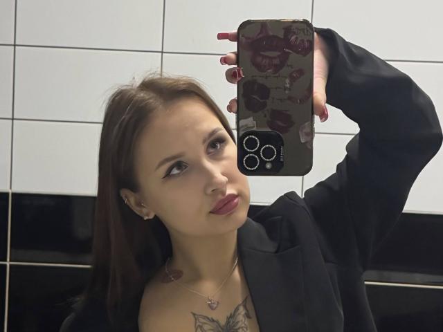 VilmaSwamy with light skin, brown hair, and pink lipstick, holding a phone with a red floral case, showing off her tits and tattoo, wearing a black shirt.