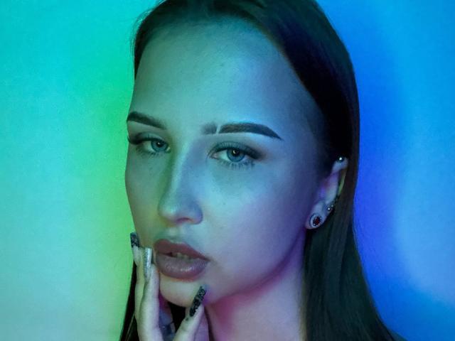 VilmaSwamy with blue-green light on face, dark brown hair, thick eyebrows, makeup, earrings, black nails, hand near chin, serious expression, blue-green gradient background.