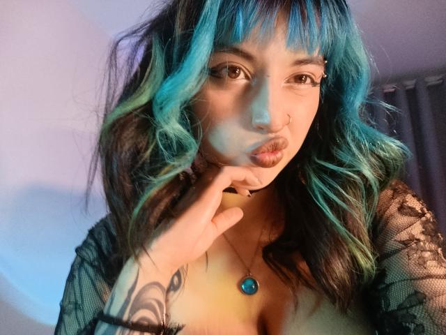 CatBleu with blue and green wavy hair, pouty lips, nose ring, black lace top, hand on chin, tattoo on wrist, blue pendant, brown eyes, soft lighting.