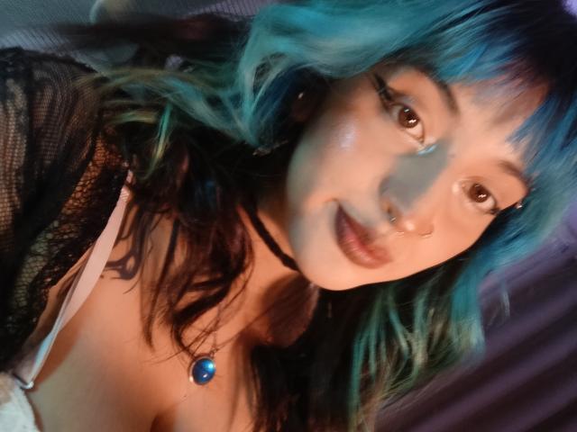 CatBleu with blue and black hair, brown eyes, nose ring, wearing a black lace top, blue pendant necklace, and a white bra, smiling softly at the camera.