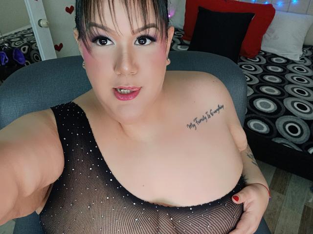 GiiannaMorrys, plus-size with large tits, wearing a sheer black top, showing a tattoo on her right shoulder, and looking at the camera with pink lipstick and pierced nose.