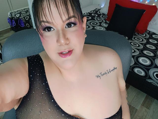 GiiannaMorrys with pink eyelids, black mesh top showing her tits, tattoo on right shoulder, taking a selfie, looking at camera, short brown hair, pink lips, light skin.