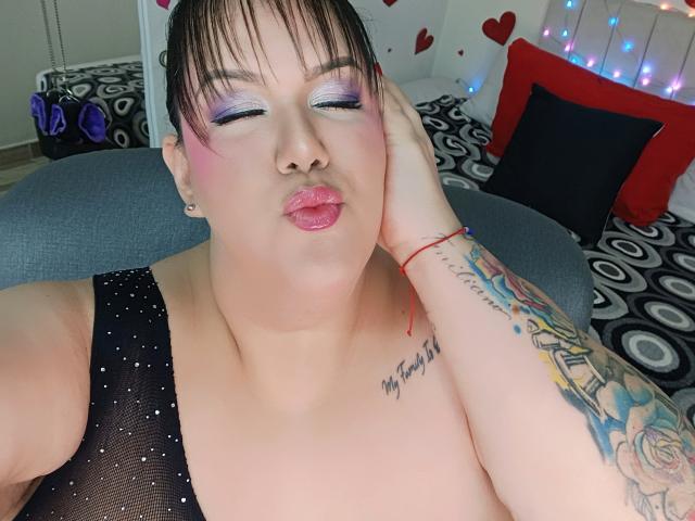 GiiannaMorrys with pink eyelids, glossy pink lips, black mesh top, tattooed arm, red bracelet, and short black hair, pouting with eyes closed. Her tits are visible.