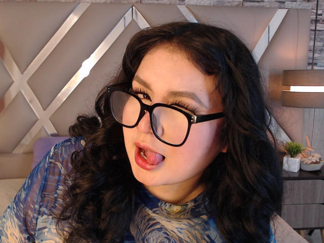 LillyMayer has curly black hair, wearing black glasses, and a blue patterned shirt. Her tongue is out, and she has long, thick eyelashes and red lipstick.