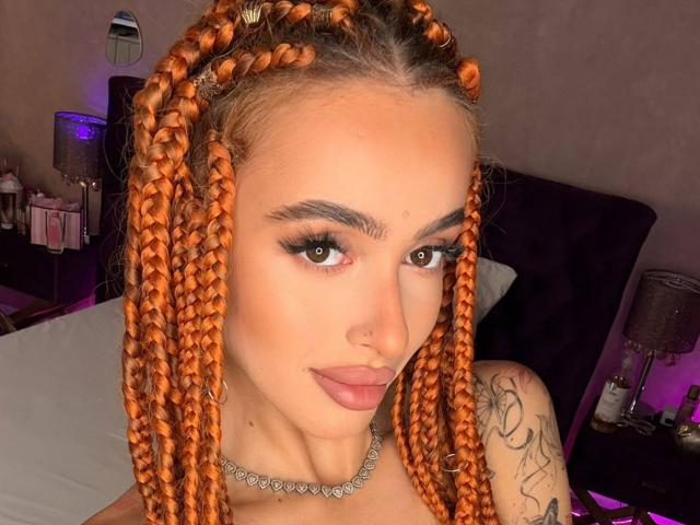 MarissaFex with orange braided hair, nose ring, tattoo on right shoulder, brown eyes, thick eyebrows, long eyelashes, pink lipstick, and a diamond necklace, in a bedroom.