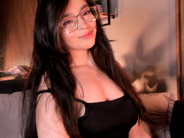 EvaMcAllister with long black hair, glasses, and a black tank top showing her tits, smiling at the camera. She's wearing a necklace and sitting on a couch.