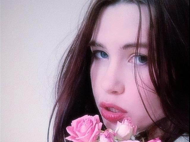 MistyRuyz with pink roses in her mouth, blue eyes, and long brown hair, looking slightly to the side. She has pale skin and pink lipstick.