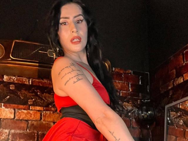 DemiMistral with long black hair, red dress, and tattoo on right arm, looking over shoulder, showing her tits and ass, in dimly lit brick-walled room.