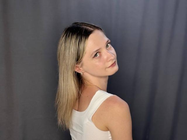 ShayneSkye, con cabello rubio, camiseta blanca, mirando por encima del hombro, con una ligera sonrisa, maquillaje sutil, fondo de cortina gris, pequeños aretes, linda, atractiva..