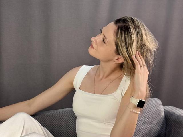 ShayneSkye, con cabello rubio, un top blanco y pantalones blancos, sentada en un sofá gris, con la mano en el cabello, mirando hacia arriba, usando un reloj inteligente y un collar delgado..