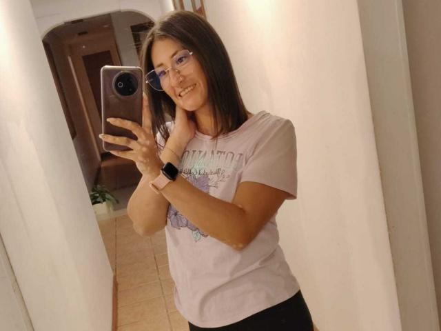 SharissWilliam with glasses, smiling, holding phone, wearing white tee, black pants, wristwatch, standing in hallway, mirror selfie.