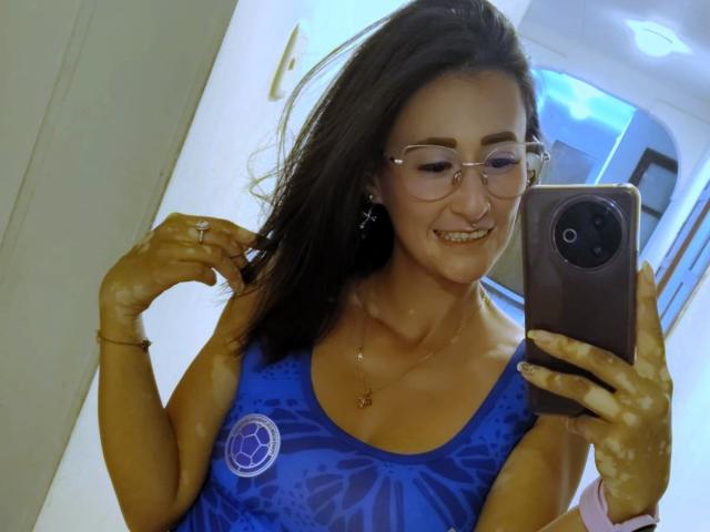 SharissWilliam with tan skin, dark brown hair, glasses, and a blue patterned tank top, smiling while taking a selfie with a smartphone. She's holding her hair with one hand.