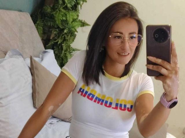 SharissWilliam with glasses, white t-shirt, yellow trim, and "MAMAJUJU" text, taking a mirror selfie, showing her tits, smiling, holding a pink phone, with a green plant.