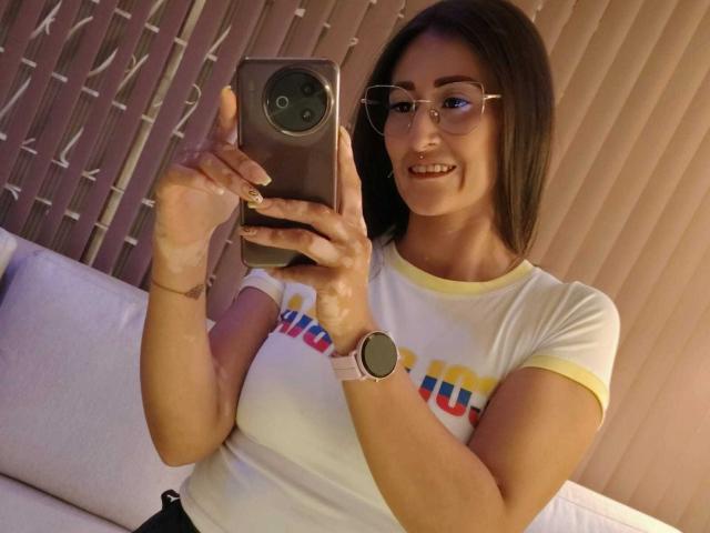 SharissWilliam with glasses, smiling, taking a selfie with a gold phone. She's wearing a white shirt with yellow trim and "UNO" text, showing her small tits and wrist tattoo.