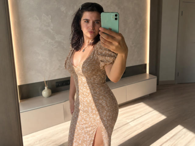 AnnaKimburli, vêtue d'une robe beige moulante à motifs floraux avec une fente sur le côté, tenant un téléphone, montrant ses courbes et ses petites seins, debout en plein soleil, regardant l'appareil photo..