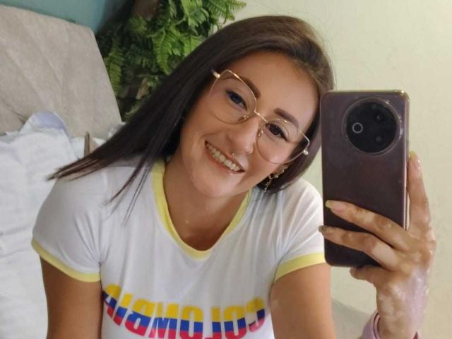 SharissWilliam with glasses, smiling, holding a phone, wearing a white shirt with yellow trim and colorful text, brown hair, nose ring, and pink wristband.