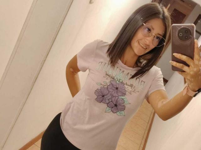 SharissWilliam with shoulder-length brown hair, glasses, smiling, wearing a pink floral T-shirt and black shorts, taking a mirror selfie with a smartphone.