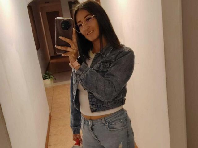 SharissWilliam with long dark hair, wearing glasses, denim jacket, white tee, and high-waisted jeans, taking a mirror selfie with a smartphone, showing a slight smile.